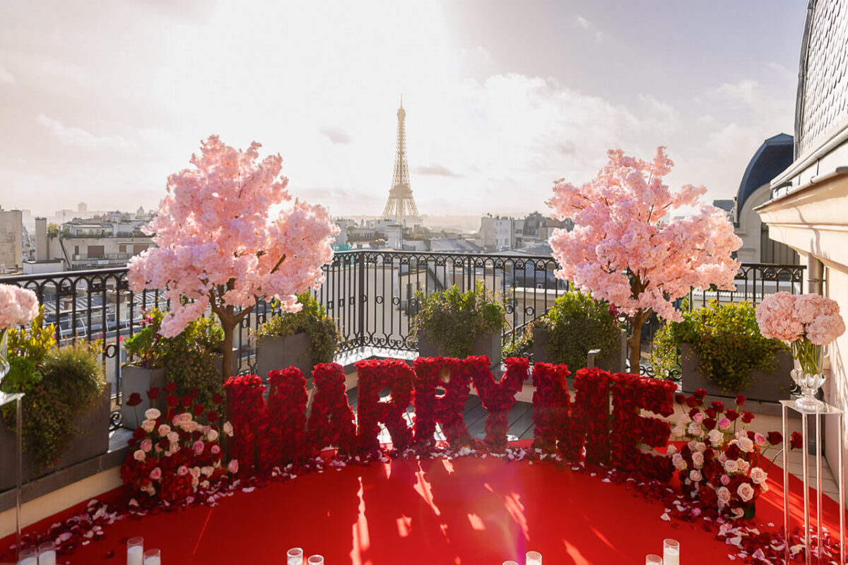 Discover our breathtaking Cherry Blossoms "Marry Me" setup on an exclusive Peninsula Paris rooftop. Big red "Marry Me" letters are flanked by cherry blossom trees and vase arrangements. A luxurious red carpet, adorned with candles and rose petals, leads to the proposal spot with the Eiffel Tower in the background, creating unmatched romance and elegance for your perfect proposal.