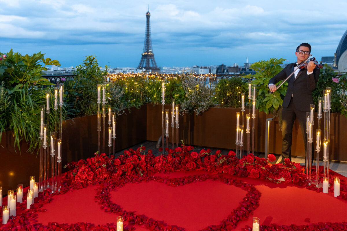 Experience the "Queen of Hearts - The Ultimate Romantic Proposal in Paris" on the exclusive Secret Table rooftop terrace of the Peninsula Hotel. This stunning setup features 250 rose petals adorning a sumptuous red carpet, creating a prominent heart marking the optimal proposal spot. Countless flameless candles, 5 towering glass candelabras, and a dazzling arrangement of silk rose flowers elevate the romantic atmosphere. With the iconic Eiffel Tower as the perfect backdrop, this luxury proposal setting is a true testament to love in the city of romance.