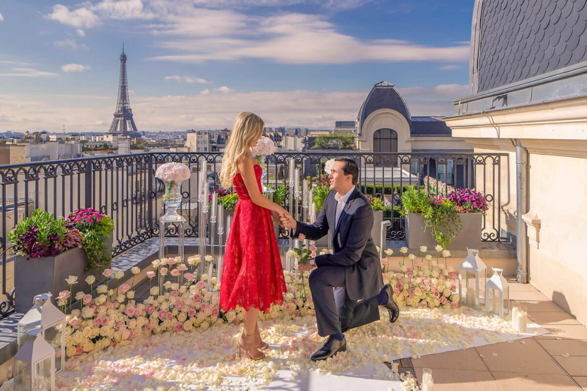 Stylish gentleman proposing on the Peninsula Secret Table Eiffel Tower terrace surrounded by 4 meters of white and pink rose bushes, scattered rose petals on a white carpet, towering candelabras, and hydrangeas in elegant transparent vases.